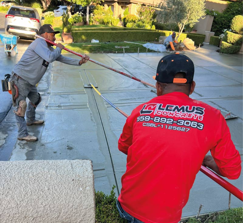 Professional concrete contractors from Lemus Concrete LLC wearing branded red long-sleeve work shirts while using bull floats to smooth a large residential driveway in Fresno.
